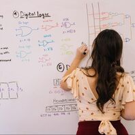 A picture of a woman, facing away from the camera, writing scientific diagrams on a whiteboard. She has long dark hair and is wearing a white and pink polka-dot top. Photo Credit: Jeswin Thomas on Unsplash 